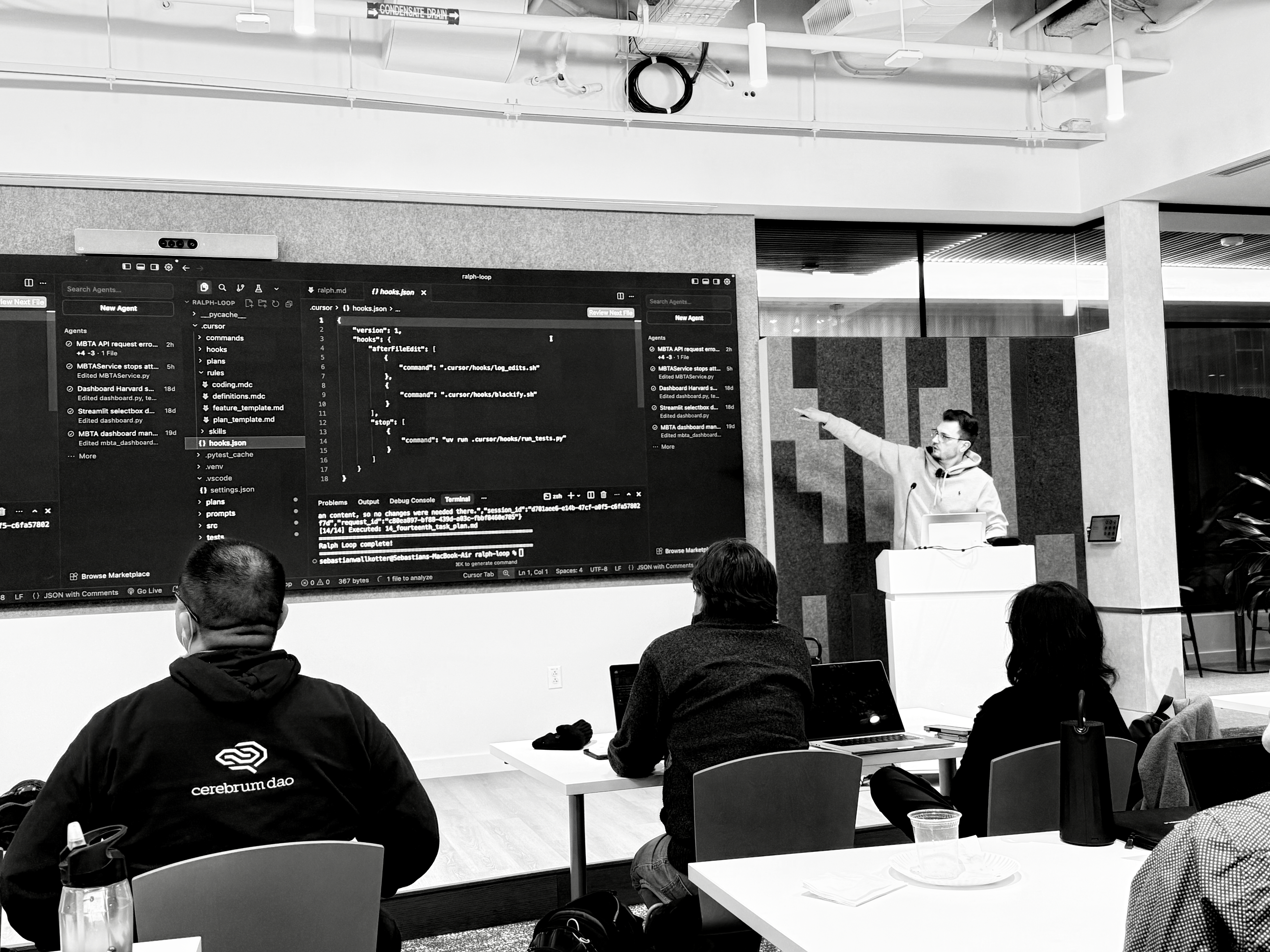 A high-contrast grayscale image captures a tech workshop. A presenter in a hoodie points animatedly at a large screen displaying lines of code. Several attendees, visible from behind, sit at tables with laptops, engrossed. The modern, bright room exudes a focused, educational atmosphere.